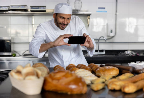 Baker taking a picture of the breads he baked Happy baker taking a picture of the breads he baked for social media using a cell phone baking Marketing and Promotion stock pictures, royalty-free photos & images