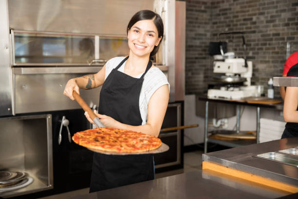 Smiling chef holding big peel with pizza Cheerful young woman working in pizza shop presenting fresh baked pizza over peel standing behind counter Baking Customer Service stock pictures, royalty-free photos & images