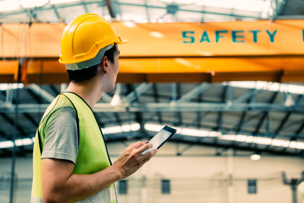 Young Asian male factory worker using a digital tablet Young Asian male heavy industral worker using a digital tablet inside manufacturing and factory site with crane behind Health and Safety in construction  stock pictures, royalty-free photos & images