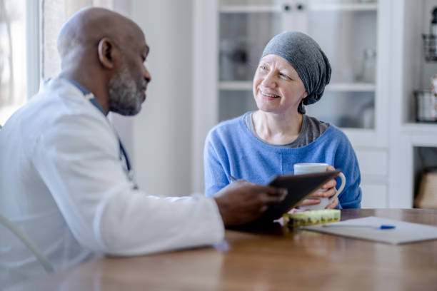 Doctor Making a Homecare Visit A male Oncologist of African decent sits with a senior woman in the comfort of her own home during a Homecare visit.  The patient is dressed comfortably and wearing a headscarf to keep her warm as the two talk about her Cancer treatment. travel insurance and cancer patients stock pictures, royalty-free photos & images