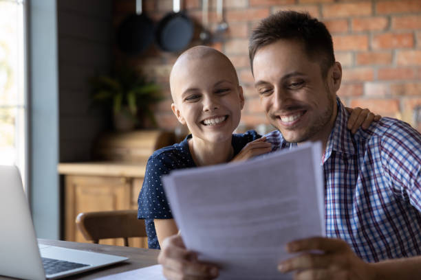 Happy wife with cancer and husband getting good news Happy wife with cancer and husband getting good news after chemotherapy, reading papers with negative oncology test result. Young married couple reviewing documents at laptop, making monthly payments travel insurance and cancer patients stock pictures, royalty-free photos & images