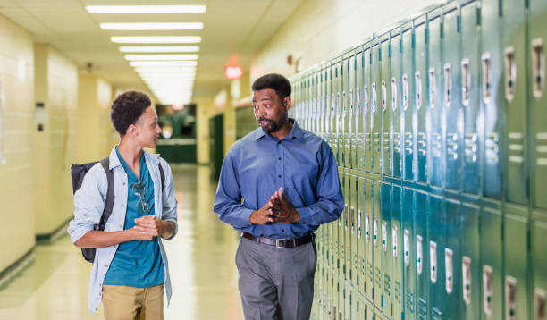 Teacher and high school student walking in hallway An African-American high school student walking with his teacher or school principal, in the hallway by a row of lockers. They are talking. The teacher has a serious expression on his face, a mature man in his 50s. The male student is an 18 year old teenage boy carrying a backpack. Inform Relevant Authorities stock pictures, royalty-free photos & images